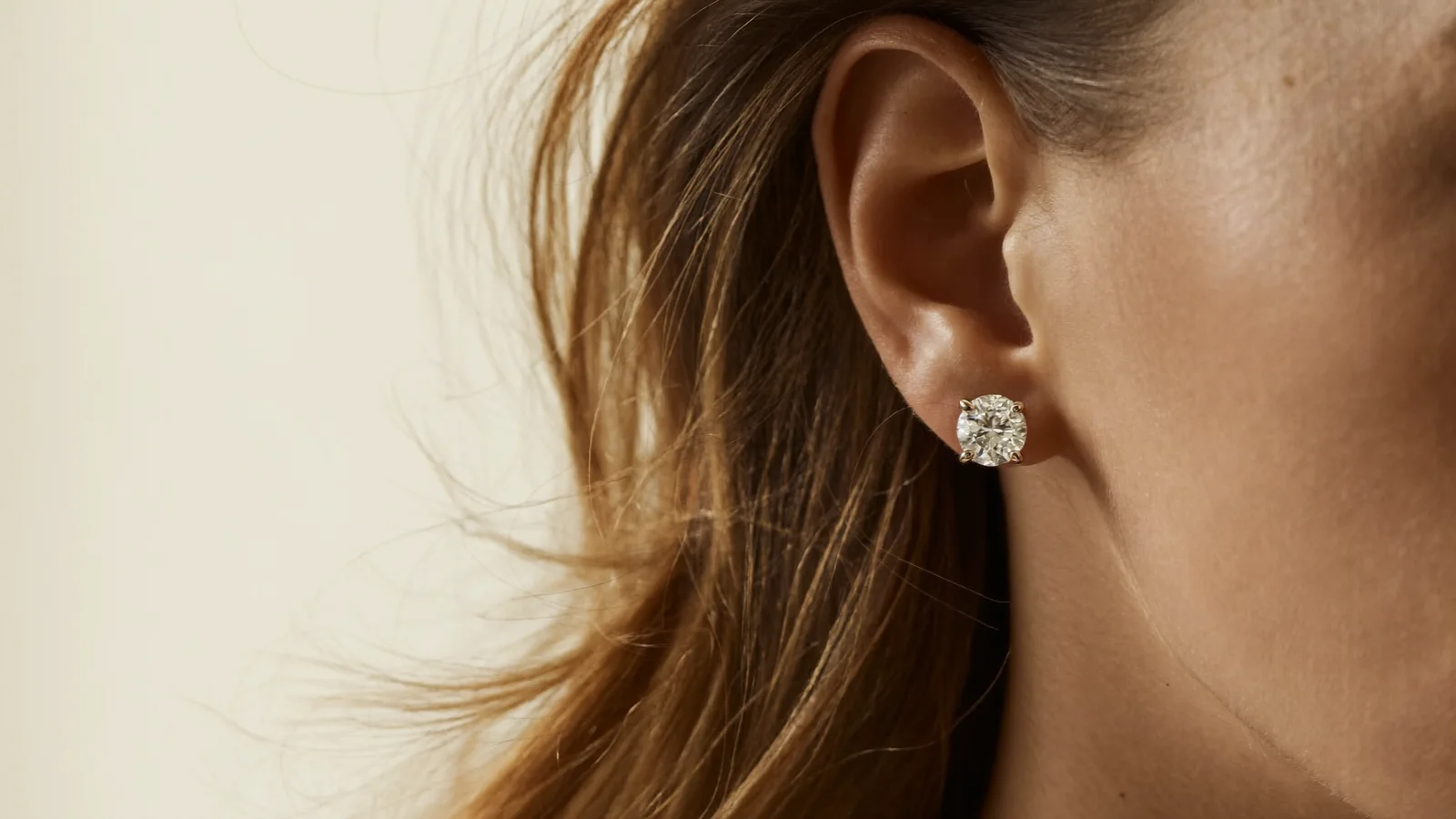 Round diamond stud earring in a four-prong setting, close-up ear profile on a cream background — DAZZ