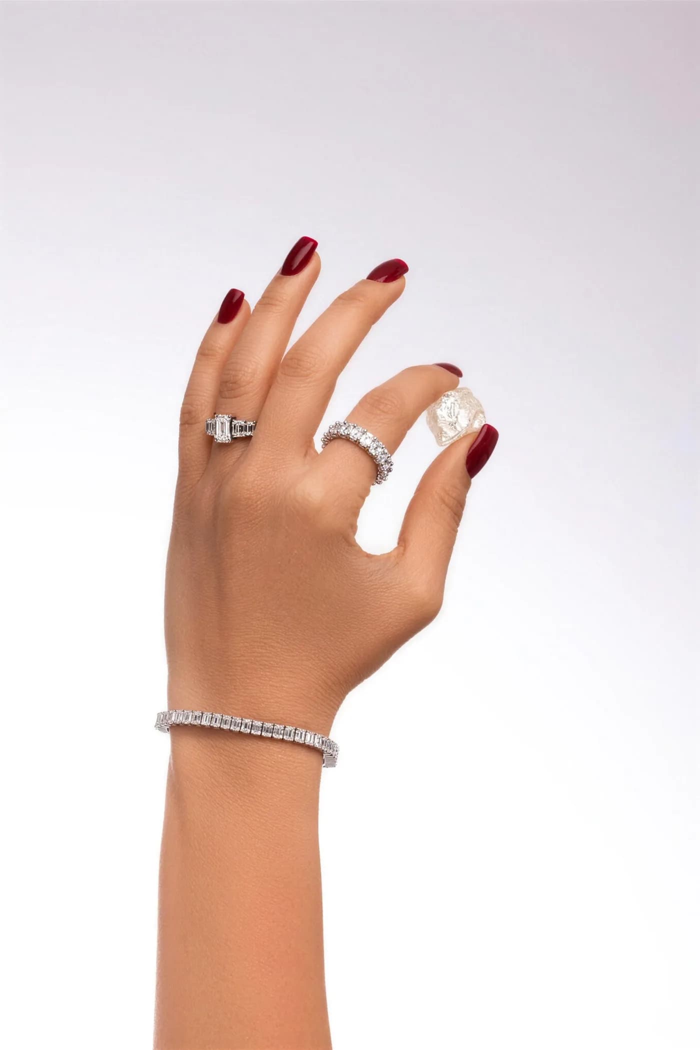 A hand wearing diamond rings and a tennis bracelet, holding a rough diamond — from the rough stone to the finished piece, DAZZ